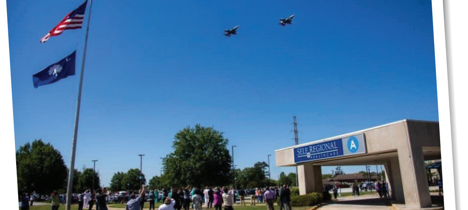 Two military F-16 jets from South Carolina Air National Guard fly over the hospital building at Self Regional Healthcare. Operation American Resolve is a salute to healthcare workers, first responders and other essential workers during COVID-19 pandemic. On left, a flagpole flies the American flag and South Carolina state flag. On right is a portico for the hospital front entrance. In center, hospital employees and first responders stand on the lawn, look up, clap and wave at the jets.